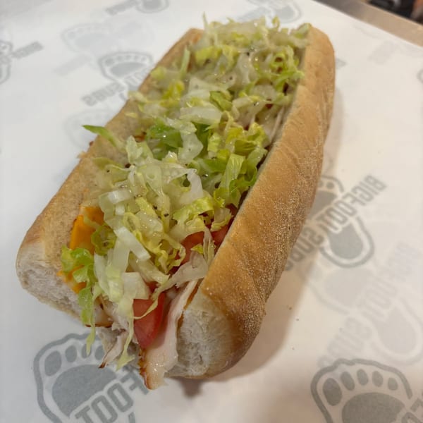 A Bigfoot Subs sandwich, filled with turkey, cheese, tomatoes, and shredded lettuce, on branded paper.